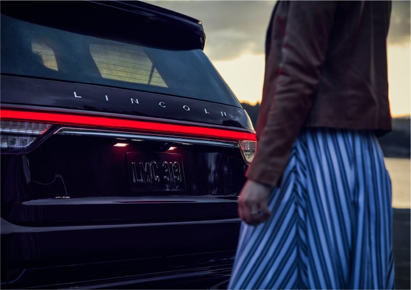 A person is shown near the rear of a 2023 Lincoln Aviator® SUV as the Lincoln Embrace illuminates the rear lights | Bill Knight Lincoln in Tulsa OK
