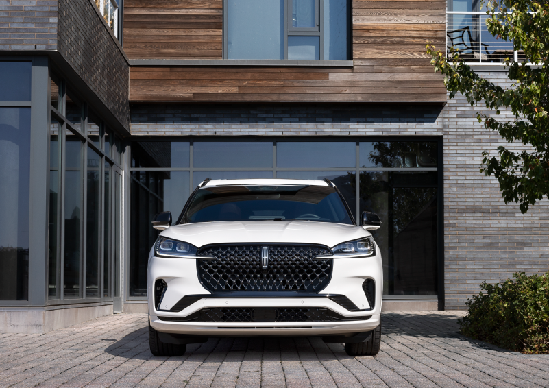 A white 2025 Lincoln Aviator® SUV is shown parked outside a modern building with gray brick and wood accents. | Bill Knight Lincoln in Tulsa OK