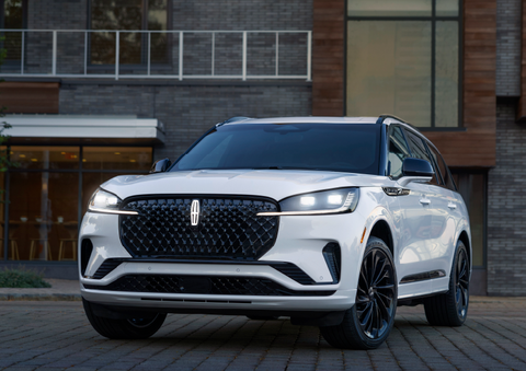 A white 2025 Lincoln Aviator® SUV is shown parked outside a modern building with gray brick and wood accents. | Bill Knight Lincoln in Tulsa OK